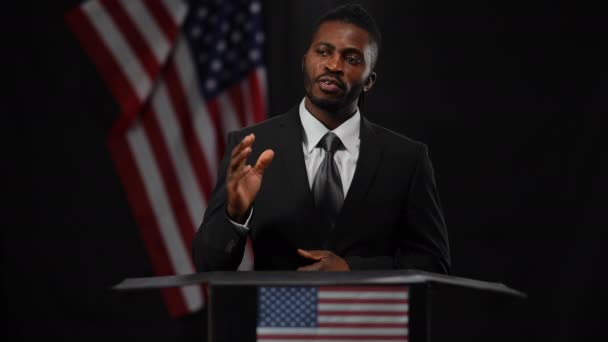 Σοβαρός επαγγελματίας Αφροαμερικάνος πολιτικός που στέκεται στο Τριμπιούν με την κάμερα να μιλάει κοιτώντας τριγύρω. Πορτρέτο του διπλωμάτη αυτοπεποίθηση απαντώντας σε ερωτήσεις με σημαία των ΗΠΑ σε μαύρο φόντο. — Αρχείο Βίντεο