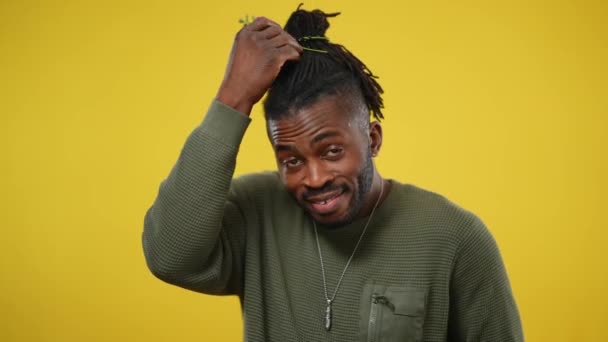 Ein positiver afroamerikanischer queerer Mann legt Blumen in Dreadlocks und lächelt in die Kamera. Fröhliche, gut aussehende LGBTQ-Person posiert vor gelbem Hintergrund. Individualitätskonzept. — Stockvideo