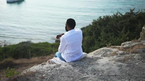Вид на расслабленного афроамериканского ЛГБТ-человека, сидящего на скале и любующегося красотой синего Средиземного моря. Счастливый беззаботный гей-турист отдыхает. Концепция спокойствия и свободы. — стоковое видео