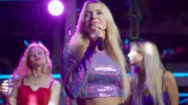 Kendine güveni tam genç bir kadının portresi. Mikrofonla şarkı söyleyen. Gece kulübünde arka planda dans eden dansçılar gibi. Başarılı beyaz şarkıcı kalabalığı eğlendiriyor. Sanat konsepti.