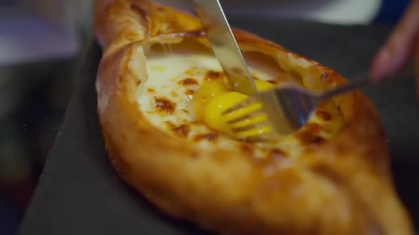 Detailní adjarianské khachapuri se syrovým vejcem na talíři. Samičí bělošské ruce s vidličkou a nožem natírajícím bílý a žloutek na pečený chutný chléb. Neznámý klient jíst gruzínské jídlo v restauraci. — Stock video