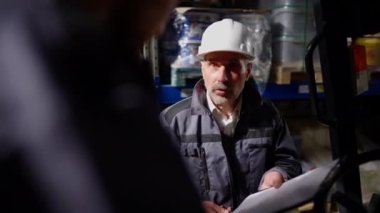 Profesyonel sakallı kıdemli nakliye acentesi depoda depo yükleyicisiyle tartışıyor ve belgelere bakıyor. İçerideki başarısızlığı yüzünden kendinden emin olmayan profesyonel bir işçi azarlıyor. Çakışma.