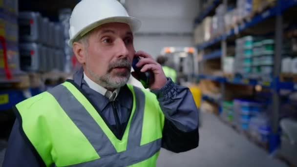 Gestionnaire d'entrepôt professionnel professionnel confiant en casque dur parlant au téléphone comme chargeur flou marchant vers un chariot élévateur à fourche en arrière-plan. Homme barbu caucasien expérimenté discutant des conditions d'expédition. — Video