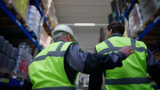 Vista posteriore di uomo anziano esperto che picchia spalla di giovane nuovo lavoratore in magazzino. Vista dell'angolo inferiore di due uomini caucasici che camminano verso il carrello elevatore all'interno parlando e sorridendo. Occupazione e sostegno. — Video Stock