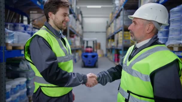 Satisfecho hombre mayor estrechando la mano con el joven empleado de almacén sonriente dentro y fuera. apretón de manos de trabajador profesional y agente de carga en el almacenamiento de la fábrica industrial. Concepto de cooperación. — Vídeos de Stock