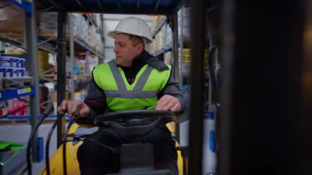 Magazijn lader rijden vorkheftruck binnen kijken rond. Vooraanzicht portret van een zelfverzekerde volwassen blanke man met een harde hoed en vest rijden voertuig in bergplaats. Live camera. — Stockvideo