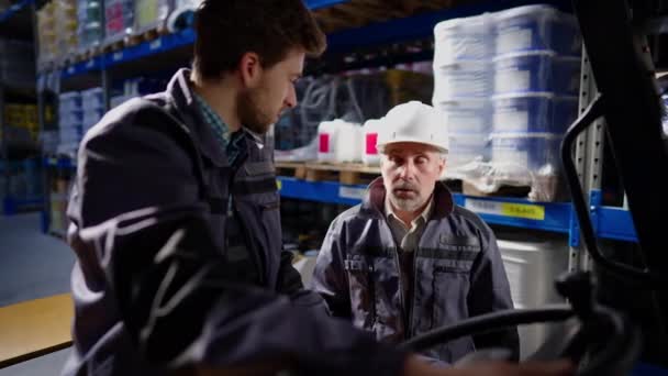 Ernstige blanke man met een witte hoed in gesprek met een jonge vorkheftruckchauffeur in een voertuig in een magazijn. Portret van ervaren professionele instructie voor nieuwe werknemer in industriële opslag. — Stockvideo