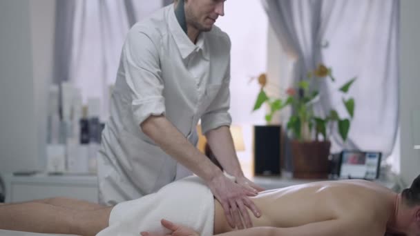 Widok z boku portret profesjonalnego masażysty masażu pleców i ramion męskiego klienta leżącego na łóżku do masażu w salonie piękności. Tatuaż młody biały mężczyzna pracuje w spa w pomieszczeniach. — Wideo stockowe