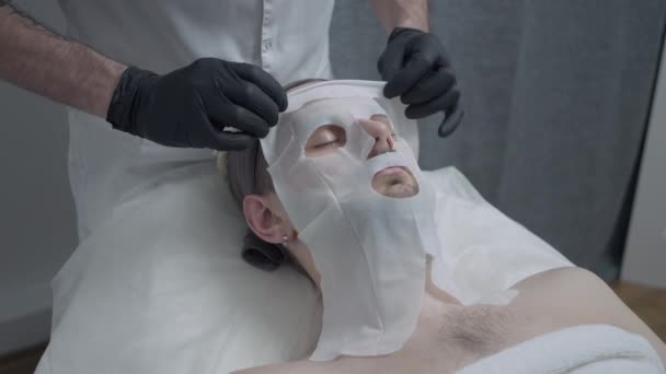 Male hands taking off cotton facial mask from face of young bearded handsome man lying with closed eyes. Unrecognizable Caucasian beautician hydrating moisturizing skin of client in spa salon. — Stock Video