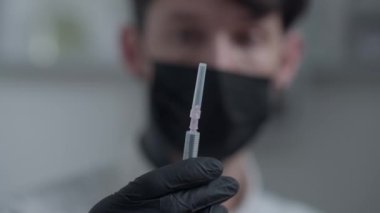 Arka planda koronavirüs maskeli bulanık bir adamın olduğu botoks dolduruculu yakın çekim şırıngası. Erkek güzellik terapisti kaplıca salonunda cilt tazelemeyi gösteriyor. Yavaş çekim.