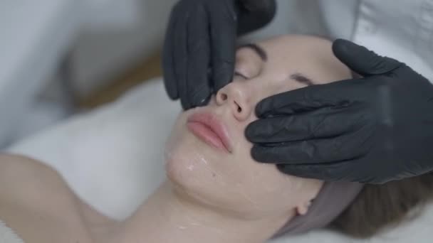 Närbild kosmetolog händer i handskar sprider ansiktspeeling på ansiktet på vackra unga kaukasiska kvinna. Underbar kvinnlig klient njuter föryngring massage i skönhetssalongen i slow motion. — Stockvideo