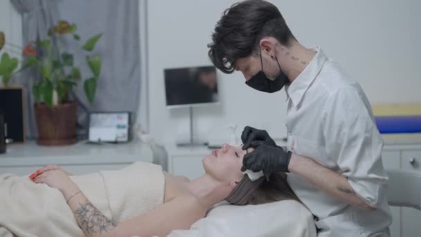 Vista lateral de la esteticista masculina en Covid-19 mascarilla limpiando la cara de la hermosa mujer acostada en el salón de spa. Joven hombre caucásico sirviendo cliente en salón de belleza en interiores en pandemia de coronavirus. Movimiento lento. — Vídeos de Stock