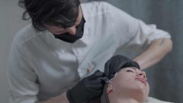 Soustředěný profesionální kosmetik podávající omlazující botoxovou náplň s injekční stříkačkou před klientem v salónu krásy. Muž v koronavirové masce obličeje dělá anti-aging proceduru pro mladou ženu. — Stock video