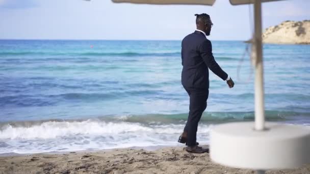 Szerokie ujęcie eleganckiego młodego Afroamerykanina spacerującego po piaszczystej plaży z turkusowymi lazurowymi falami morskimi toczącymi się na wybrzeżu w zwolnionym tempie. Malowniczy śródziemnomorski krajobraz morski, spokojny biznesmen spacerujący — Wideo stockowe