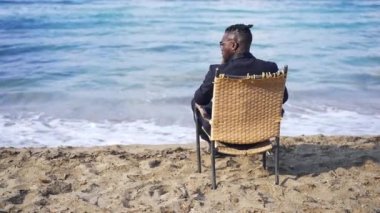 Arkadaki hasır koltukta resmi takım elbiseli Afro-Amerikan genç adam oturmuş mavi dalgaların güzelliğini seyrediyor ağır çekimde yuvarlanıyor. Rahatlamış kendine güvenen turist kumsalda tatilin tadını çıkarıyor..