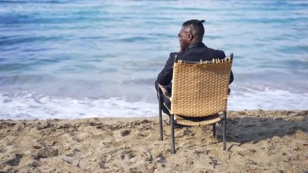 Back view wicker chair with African American young man in formal suit sitting admiring beauty of azure blue waves rolling in slow motion. Relaxed confident tourist enjoying vacations on sandy beach. — Stock Video
