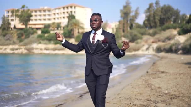 Portrait de joyeux heureux marié afro-américain heureux en costume élégant dansant marchant sur la plage de sable fin et ensoleillée de la Méditerranée souriant. Joyeux bel homme qui s'amuse le jour du mariage en plein air. — Video