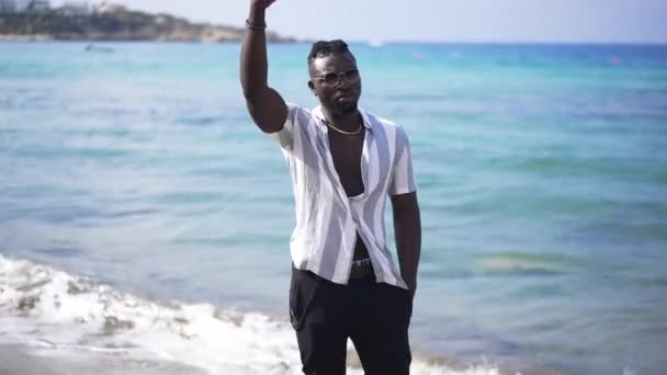 Portrait of positive confident handsome African American man in sunglasses waving inviting someone standing on sunny sandy beach at resort. Carefree tourist enjoying vacations on Mediterranean island. — Stock Video