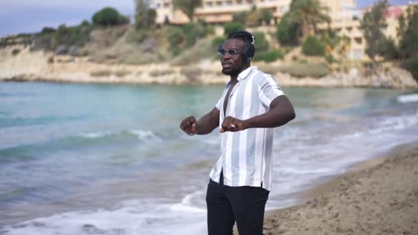 Kumlu plajda müzik eşliğinde kulaklıkla dans eden mutlu, rahat erkek Afrikalı Amerikalı turistin portresi. Kulaklıklı neşeli yakışıklı adam Akdeniz kıyısında dinleniyor.. — Stok video