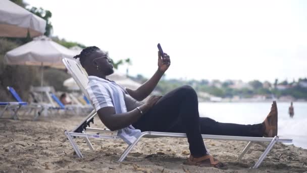 Боковой вид позитивного афроамериканского туриста-мужчины, использующего мобильное приложение для видеочата, лежащего на шезлонге на песчаном пляже. Широкий снимок расслабленного красивого молодого человека, говорящего жестом в замедленной съемке. — стоковое видео