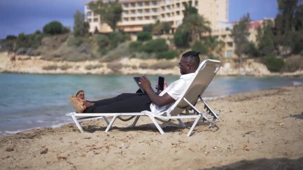 砂のビーチで日なたぼっこをしているスマートフォンのタブレットのテキストメッセージでインターネットサーフィンの忙しいアフリカ系アメリカ人男性。観光地からオンラインで働く男性フリーランスの集中。スローモーション. — ストック動画