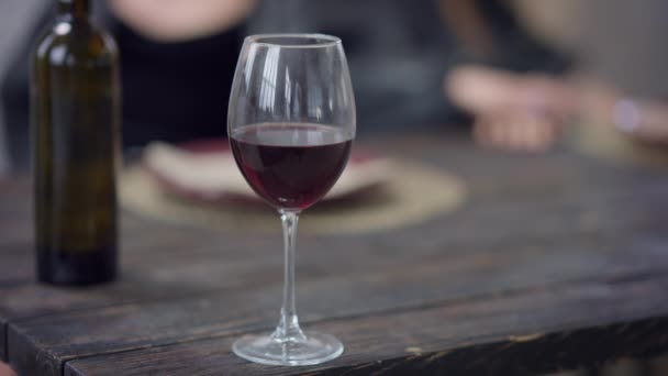 Rotwein in Großaufnahme im Glas auf einem Holztisch mit verschwommenen kaukasischen Händen im Hintergrund. Drinnen wird Alkohol getrunken. Zeitlupe. — Stockvideo