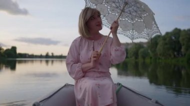 Mutlu ikili olmayan trans kadın beyaz güneş şemsiyesini yavaş çekimde döndürüyor ve kameraya bakıp gülümsüyor. Gölde teknede poz veren gevşemiş beyaz kadın portresi..
