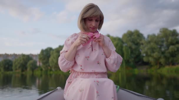 Mulher trans romântico em vestido elegante amarrando lenço rosa no pescoço sentado em barco no lago. Retrato de pessoa elegante caucasiano gênero-fluido descansando namoro ao ar livre na noite de verão. — Vídeo de Stock