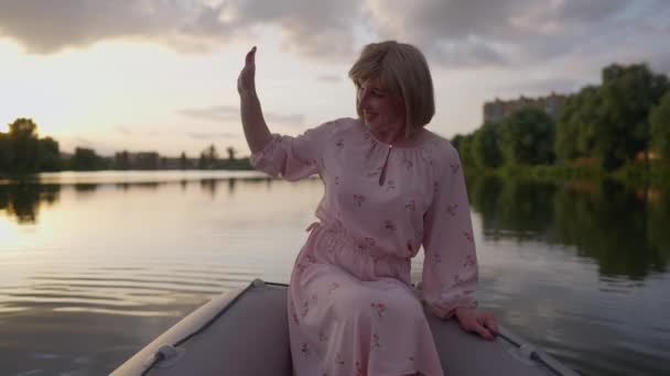 Feliz mujer trans encantadora saludando invitando a alguien sentado en el barco en el lago de verano. Retrato de una persona caucásica positiva y confiada que descansa al atardecer al aire libre. — Vídeos de Stock