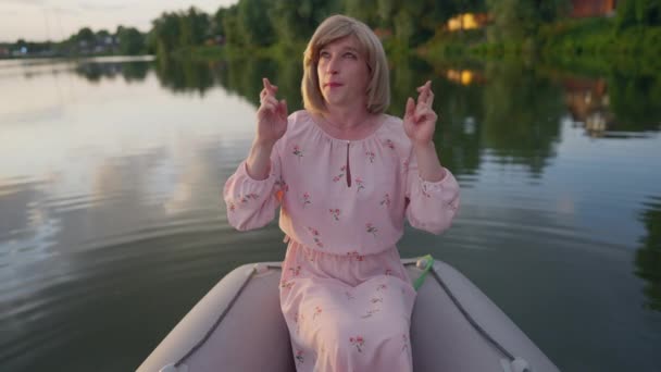 Femme trans positive croisant les doigts fermant les yeux faisant un souhait assis dans le bateau sur le lac. Portrait de heureux détendu Caucasien non-binaire souhaitant à l'extérieur au coucher du soleil. Espérance et identité de genre. — Video
