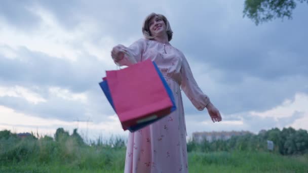 Возбужденная счастливая трансгендерная женщина поднимает руки, а сумки с покупками крутятся на улице в замедленной съемке. Портрет веселого кавказского стильного покупателя гендерной жидкости на летнем лугу. — стоковое видео
