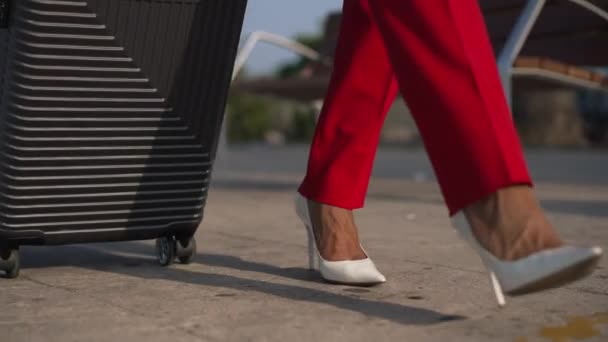 La cámara en vivo sigue a las piernas femeninas en pantalones rojos y tacones altos blancos caminando con equipaje al aire libre en cámara lenta. Una mujer de negocios irreconocible en viaje de negocios. Concepto de llegada y salida. — Vídeo de stock