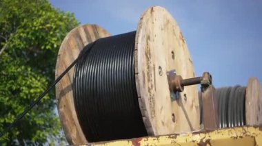 Arka planda mavi gökyüzü olan büyük bir elektrik teli açık havada yuvarlanıyor. İnşaat alanında gün ışığında dönen endüstriyel ekipmanlar.