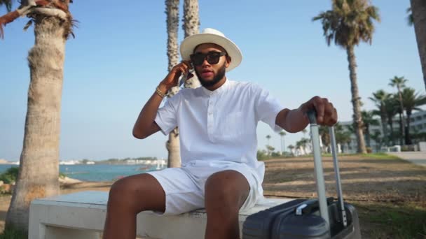 Handsome positive African American man sitting on summer resort with travel bag talking on the phone. Portrait of happy male tourist with luggage with picturesque Mediterranean landscape at background — Stock Video