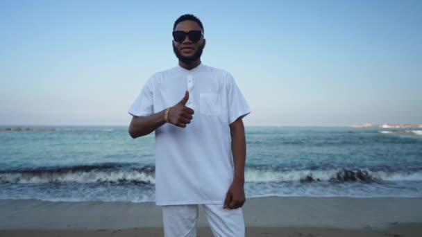 Retrato de um homem afro-americano confiante em óculos de sol mostrando o polegar em pé na praia de areia com ondas azul-turquesa do mar Mediterrâneo rolando em câmera lenta na costa. Turismo e lazer. — Vídeo de Stock