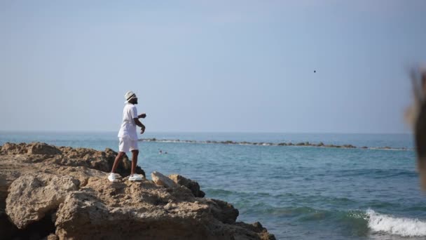 Amplio plano de jóvenes millennials afroamericanos masculinos lanzando piedras en espumoso azul del mar Mediterráneo en cámara lenta. Hombre desesperado frustrado en el acantilado en un paisaje pintoresco. — Vídeos de Stock