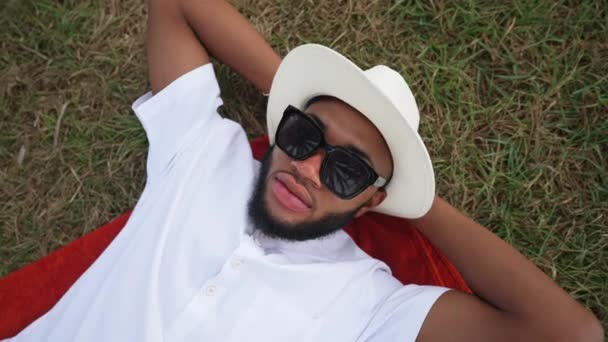 Widok z góry beztroski przystojny mężczyzna w białej koszuli polo kapelusz i okulary przeciwsłoneczne leżące na zielonej trawie na zewnątrz. Zrelaksowany Afroamerykanin pewny siebie turysta odpoczywający na letnim trawniku w kurorcie. — Wideo stockowe