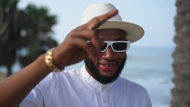 Primer plano del alegre hombre afroamericano feliz en gafas de sol saludando mirando a la cámara sonriendo con el mar azul borroso y la palmera al fondo. Turista masculino posando al aire libre en el resort Mediterráneo. — Vídeo de stock