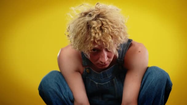 Portrait of Caucasian androgynous LGBT man sitting on hunkers looking at camera. Confident serious queer person posing at yellow background in slow motion. — Stock Video