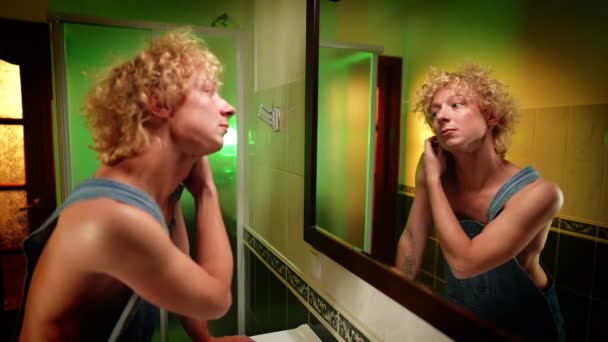 Männliche kaukasische queere Person, die Spiegelungen im Spiegel im Badezimmer bewundert. Porträt eines androgynen, gut aussehenden jungen LGBT-Mannes zu Hause im Haus. Selbstbewusstsein und Individualität. — Stockvideo