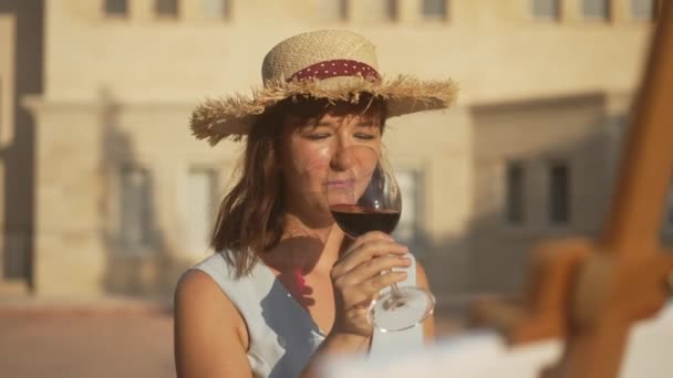 Young positive Caucasian woman drinking red wine in slow motion admiring picture on easel outdoors. Portrait of happy beautiful painter looking at artwork in sunlight enjoying beverage. Art concept. — Stock Video