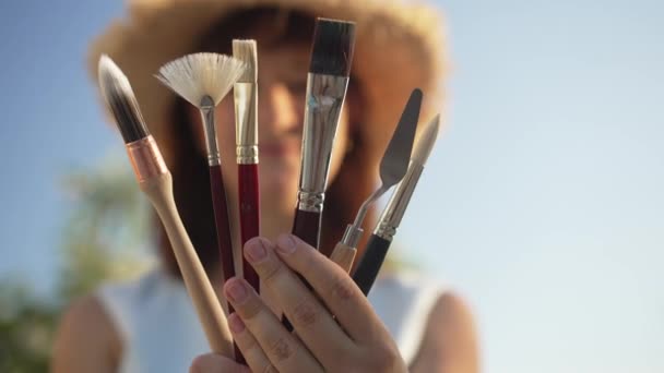在明亮的蓝天背景下，白种人女性手中的特写工具与模糊的天才快乐女人在一起。有创意的女性画家在室外展示画笔和刀具. — 图库视频影像