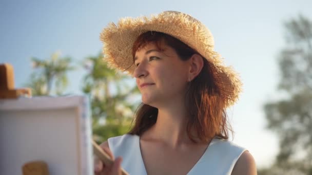 Happy confiante pintor feminino sorrindo fazendo traços na tela com escova e olhando para longe. Retrato de mulher bonita caucasiana talentosa em pintura chapéu de palha com céu azul de verão no fundo. — Vídeo de Stock