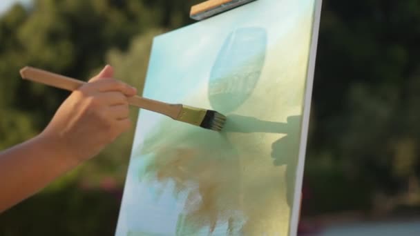 Close-up vrouwelijke hand schilderen op doek met silhouet schaduw van wijnglas reflecteren in de zon. Onherkenbare blanke vrouw genieten van hobby buiten op zonnige zomerdag. Langzame beweging. — Stockvideo