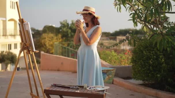 Retrato de mulher criativa bebendo chá da tarde admirando quadro no cavalete ao ar livre. Feliz talentosa artista feminina caucasiana apreciando a pintura no dia ensolarado de verão. Criatividade e conceito de arte. — Vídeo de Stock