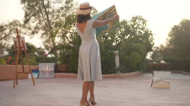 Amplio plano de feliz orgullosa pintora caucásica bailando con cuadro de paisaje en cámara lenta. Retrato de mujer delgada elegante talentosa alegre disfrutando de estilo de vida y pasatiempo al atardecer dorado. — Vídeo de stock