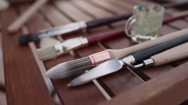 Gros plan pinceaux et couteaux à peindre sur table en bois avec main blanche femelle prenant pinceau au ralenti laissant. Femme créative talentueuse et méconnaissable peignant à l'extérieur. — Video