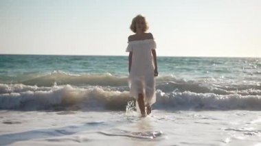 Beyaz elbiseli, beyaz tenli, muhteşem beyaz bir kadının ön manzara portresi kumlu sahilde ağır çekimde, köpüklü mavi Akdeniz dalgalarıyla yürüyor. Kendine güvenen bir kadın gün ışığında geziniyor..