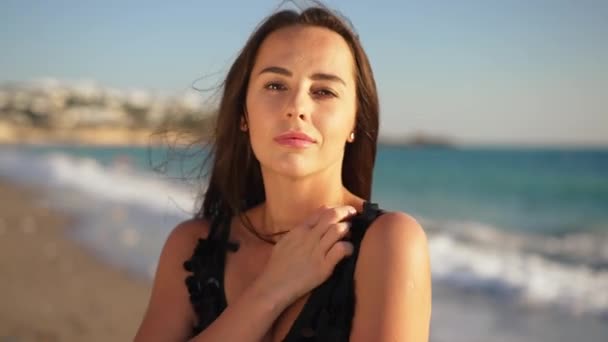 Retrato de bela mulher caucasiana confiante em vestido preto posando à luz do sol na praia de areia. Vista frontal da jovem senhora magro olhando para a câmera com turquesa ondas do mar Mediterrâneo no fundo. — Vídeo de Stock