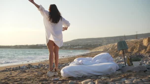 Vista posterior de la relajada mujer joven y delgada feliz que se extiende bebiendo café de la mañana de pie en la playa de arena del mar Mediterráneo con cama blanca acolchada. Concepto de relajación y confort. Movimiento lento. — Vídeo de stock